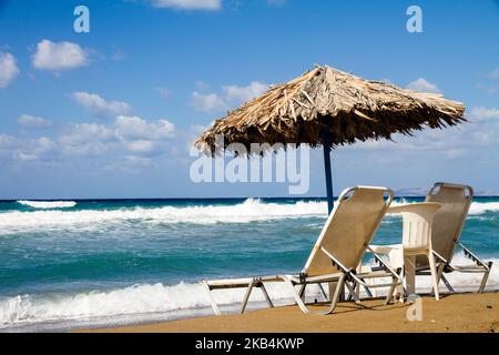 Due sedie posizionato su di una spiaggia di sabbia unset paglia ombrellone, nella parte anteriore di mare mosso con onde alte e blu cielo nuvoloso Foto Stock