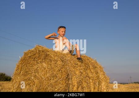 Al tramonto il ragazzo siede sul campo per una balla di paglia Foto Stock