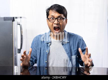 Ritratto di un giovane maschio indiano di mezza età che indossa occhiali seduti a tavola con faccia arrabbiata su sfondo bianco a casa Foto Stock