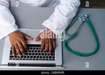 Primo piano vista dall'alto di un medico africano maschio irriconoscibile in uniforme cappotto bianco che lavora digitando sul computer portatile seduto alla scrivania con Foto Stock