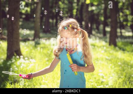 adorabile ragazza bionda che si diverte con le bolle di sapone all'aperto in verde foresta di primavera Foto Stock