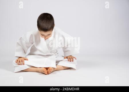 Un ragazzino in un kimono pratica il karate, siede in una posa a farfalla e pratica lo stretching, un posto per il testo. Foto Stock