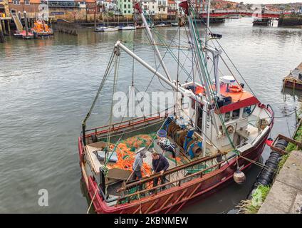 La poppa di un peschereccio con reti a strascico sul ponte. Due pescatori assistono ad un problema con una catena. Il porto è oltre. Foto Stock