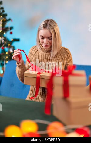 Una donna in un maglione celebra il nuovo anno e apre un regalo trovato sotto l'albero. Concetto di vacanza. La ragazza disimballa un regalo di Natale. Primo piano Foto Stock