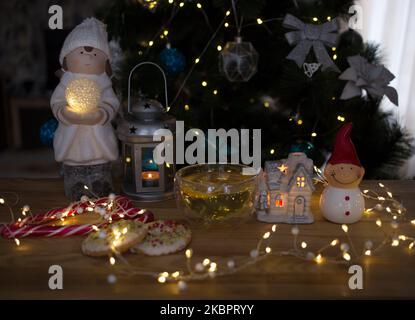 Accogliente foto d'atmosfera su un tema natalizio. Una tazza trasparente con tè, un riflesso delle luci di un ghirlaio in esso contenuto. Pupazzo di neve ricordo, biscotti, lan Foto Stock