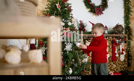 Il bambino decora un albero di Natale per Natale. Il ragazzo appende un giocattolo di Capodanno sull'albero di Natale. Appendere decorazione di Natale. Foto Stock