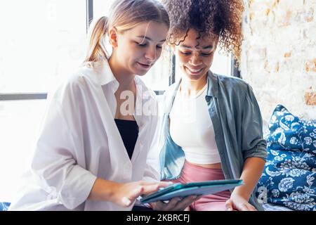 Due studenti di sesso femminile multietnica seduti al tavolo con un computer tablet, che parlano tra loro e si divertono. Istruzione, scuola superiore, persone e tec Foto Stock