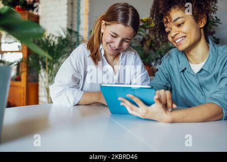 Due studenti di sesso femminile multietnica seduti al tavolo con un computer tablet, che parlano tra loro e si divertono. Istruzione, scuola superiore, persone e tec Foto Stock