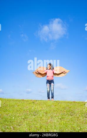 colpo verticale, ragazza felice eccitata con ali artificiali che ballano come un uccello sulla collina in verde prato contro il cielo blu - concetto di libertà Foto Stock