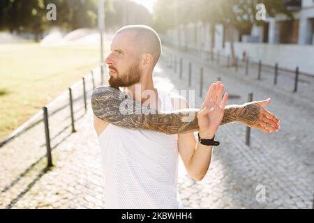 Giovane uomo tatuato muscoloso che fa esercizi, stretching presto al mattino, calvo bearded ragazzo in sportswear stretching braccia prima di jogging Foto Stock