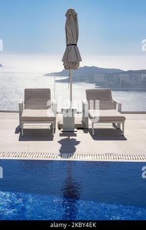 Piscina in un hotel resort a 5 stelle con sedie a sdraio e asciugamano che si affaccia su una splendida vista sulla Caldera sul mare Egeo con tradizionale architettura a terrazze greca a calce e mediterranea di case e resort. Isola di Santorini nelle Cicladi, Grecia durante una calda giornata estiva di sole. Santorini è di solito sovraffollata in quanto è famosa per il suo tramonto, che attrae soprattutto coppie da tutto il mondo per la magica vista del tramonto sul vulcano, Ma quest'anno a causa della pandemia di Coronavirus Covid-19 ci sono meno arrivi del solito, riducendo anche il reddito del PIL del conteggio Foto Stock