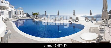 Piscina in un hotel resort a 5 stelle con sedie a sdraio e asciugamano che si affaccia su una splendida vista sulla Caldera sul mare Egeo con tradizionale architettura a terrazze greca a calce e mediterranea di case e resort. Isola di Santorini nelle Cicladi, Grecia durante una calda giornata estiva di sole. Santorini è di solito sovraffollata in quanto è famosa per il suo tramonto, che attrae soprattutto coppie da tutto il mondo per la magica vista del tramonto sul vulcano, Ma quest'anno a causa della pandemia di Coronavirus Covid-19 ci sono meno arrivi del solito, riducendo anche il reddito del PIL del conteggio Foto Stock