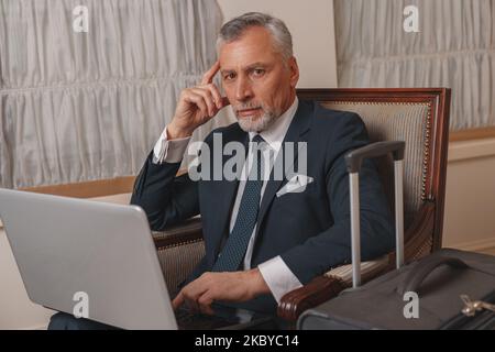 Uomo d'affari che lavora sul computer portatile mentre si siede nella lobby dell'hotel e attende il check-in Foto Stock