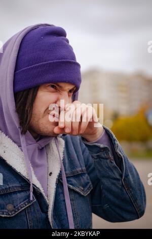 Ritratto di un uomo che si annida al freddo, in piedi sulla strada cittadina. Ragazzo congelato in abiti caldi con il muso, il naso che puliva Foto Stock
