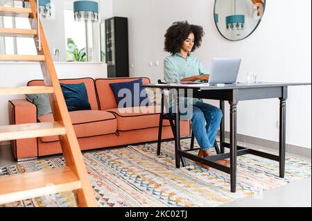 Giovane donna d'affari multirazziale concentrata in abbigliamento casual seduto alla scrivania in casa ufficio, lavorando in remoto sul notebook. Donna sorridente freelancer che scrive, rispondendo alle e-mail Foto Stock