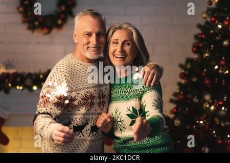 coppia matura felice in maglioni di festa che tengono scintillanti luccicanti la vigilia di natale nel paese Foto Stock