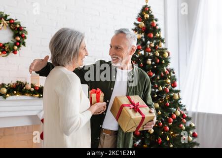 donna matura allegra scambiando regali di natale con marito felice vicino abete decorato Foto Stock