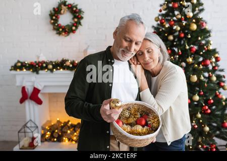 allegra donna di mezza età appoggiata al marito con cestino di vimini che guarda la palla di natale Foto Stock