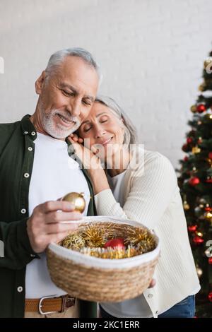 smiling middle aged woman leaning on husband with wicker basket near christmas tree Stock Photo