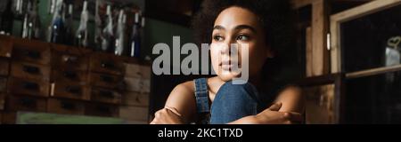 giovane e sognante donna afroamericana seduta in laboratorio d'arte e guardando via, banner Foto Stock