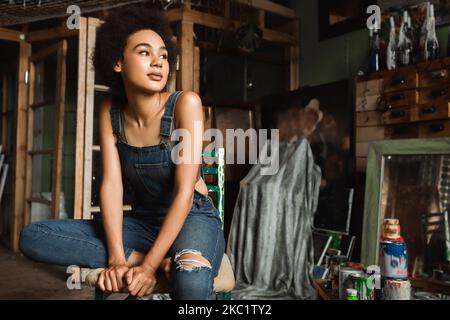 young african american woman in denim overalls looking away in art workshop near cans with paint Stock Photo