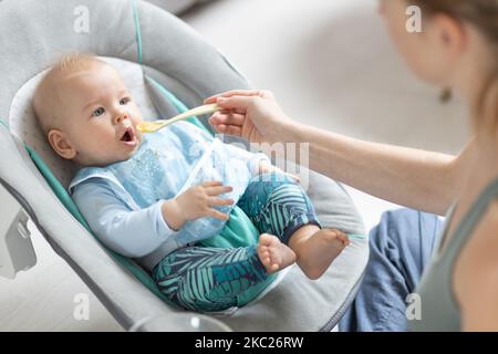 Il cucchiaio della madre che alimenta il bambino del suo bambino del bambino nella sedia del bambino con purea di frutta. Concetto di introduzione degli alimenti solidi per bambini Foto Stock