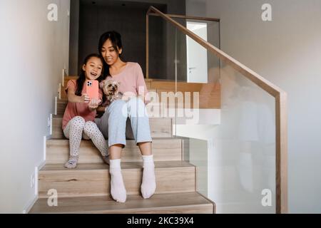 Madre e figlia asiatica che prendono selfie mentre si siedono con il loro cane sulle scale a casa Foto Stock