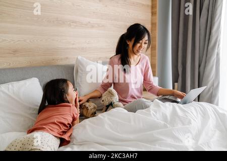 Madre e figlia asiatica che usano un computer portatile mentre si siedono a letto a casa Foto Stock