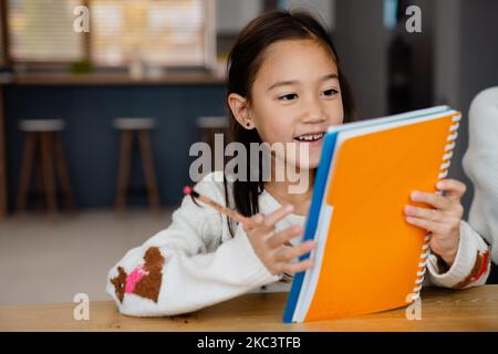 Ragazza asiatica sorridente e facendo i compiti mentre si siede al tavolo a casa Foto Stock