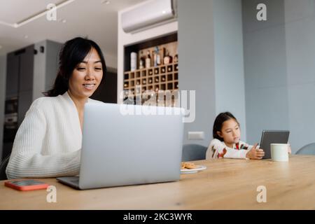 Donna asiatica e sua figlia utilizzando gadget mentre si siede al tavolo a casa Foto Stock