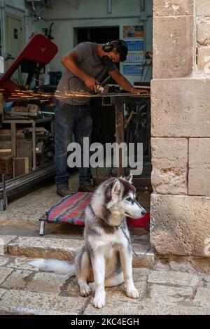 Un artigiano lavora nella sua piccola fabbrica utilizzando una smerigliatrice angolare con scintille che volano mentre il suo cane Husky mantiene la guardia. Foto Stock