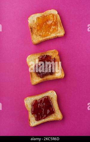 Tre fette croccanti di pane tostato con fragola, albicocca e confettura di fichi su sfondo rosa brillante Foto Stock