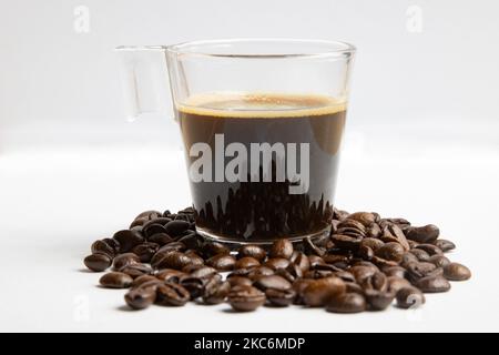 Una tazza di caffè di vetro con un espresso nella tazza accanto alla varietà di chicchi di caffè arabica organici crudi su sfondo bianco isolato. Amsterdam, Paesi Bassi il 30 dicembre 2020 (Foto di Nicolas Economou/NurPhoto) Foto Stock