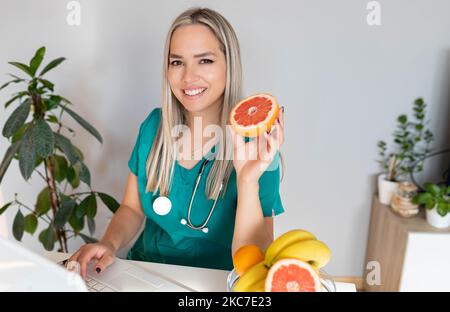 Nutrizionista femminile con frutta che lavora alla sua scrivania. Nutrizionista sorridente nel suo ufficio, mostra verdura e frutta sane, assistenza sanitaria Foto Stock