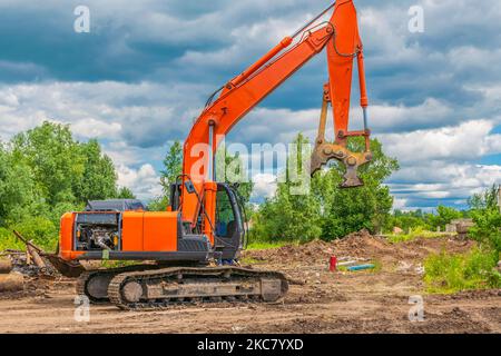 Il grande escavatore cingolato arancione con carro di frantumazione si trova di fronte al campo agricolo con alberi e cielo estivo nuvoloso e blu. Foto Stock