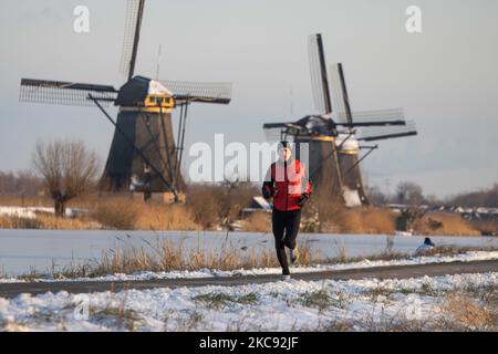 Un uomo con giacca rossa come visto correre nella neve a Kinderdijk. Una magica scriglia intorno al tramonto e crepuscolo con il cielo limpido d'inverno ai canali ghiacciati e ai mulini a vento di Kinderdijk nei Paesi Bassi. Il paese sta affrontando il clima artico, le temperature basse severe, le nevicate ed i venti alti dovuto la tempesta Darcy che ha causato i problemi nei trasporti e nella vita di tutti i giorni. Molti dei canali sono stati ghiacciati perché la temperatura è inferiore a zero a causa del clima freddo che a volte raggiunge i -16C, mentre il terreno è coperto di neve. Kinderdijk e i mulini a vento nel polder Alblasserwaard, è un'attrazione famosa Foto Stock