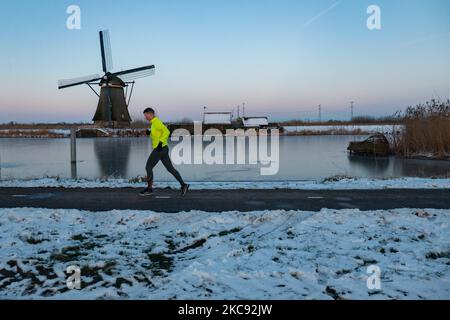 Un uomo come visto che corre davanti ad un mulino a vento. Una magica scriglia intorno al tramonto e crepuscolo con il cielo limpido d'inverno ai canali ghiacciati e ai mulini a vento di Kinderdijk nei Paesi Bassi. Il paese sta affrontando il clima artico, le temperature basse severe, le nevicate ed i venti alti dovuto la tempesta Darcy che ha causato i problemi nei trasporti e nella vita di tutti i giorni. Molti dei canali sono stati ghiacciati perché la temperatura è inferiore a zero a causa del clima freddo che a volte raggiunge i -16C, mentre il terreno è coperto di neve. Kinderdijk e i mulini a vento nel polder Alblasserwaard, è una famosa attrazione, un landmar olandese Foto Stock