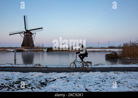 Un uomo come visto in bicicletta la sua mini bici nella pista ciclabile davanti a un mulino a vento. Una magica scriglia intorno al tramonto e crepuscolo con il cielo limpido d'inverno ai canali ghiacciati e ai mulini a vento di Kinderdijk nei Paesi Bassi. Il paese sta affrontando il clima artico, le temperature basse severe, le nevicate ed i venti alti dovuto la tempesta Darcy che ha causato i problemi nei trasporti e nella vita di tutti i giorni. Molti dei canali sono stati ghiacciati perché la temperatura è inferiore a zero a causa del clima freddo che a volte raggiunge i -16C, mentre il terreno è coperto di neve. Kinderdijk e i mulini a vento nel polder Alblasserwaard, è una truffa Foto Stock