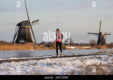 Un uomo con giacca rossa come visto correre nella neve a Kinderdijk. Una magica scriglia intorno al tramonto e crepuscolo con il cielo limpido d'inverno ai canali ghiacciati e ai mulini a vento di Kinderdijk nei Paesi Bassi. Il paese sta affrontando il clima artico, le temperature basse severe, le nevicate ed i venti alti dovuto la tempesta Darcy che ha causato i problemi nei trasporti e nella vita di tutti i giorni. Molti dei canali sono stati ghiacciati perché la temperatura è inferiore a zero a causa del clima freddo che a volte raggiunge i -16C, mentre il terreno è coperto di neve. Kinderdijk e i mulini a vento nel polder Alblasserwaard, è un'attrazione famosa Foto Stock