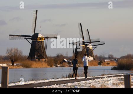 Una coppia come visto in esecuzione a Kinderdijk. Una magica scriglia intorno al tramonto e crepuscolo con il cielo limpido d'inverno ai canali ghiacciati e ai mulini a vento di Kinderdijk nei Paesi Bassi. Il paese sta affrontando il clima artico, le temperature basse severe, le nevicate ed i venti alti dovuto la tempesta Darcy che ha causato i problemi nei trasporti e nella vita di tutti i giorni. Molti dei canali sono stati ghiacciati perché la temperatura è inferiore a zero a causa del clima freddo che a volte raggiunge i -16C, mentre il terreno è coperto di neve. Kinderdijk e i mulini a vento nel polder Alblasserwaard, è una famosa attrazione, un punto di riferimento olandese e. Foto Stock