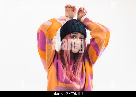 Divertente ritratto in studio di ragazza adolescente caucasica dai capelli rosa in una beanie nera e cardigan colorato facendo orecchie di coniglio mettendo le mani sopra la testa. Scatto di primo piano medio. Sfondo bianco. Foto di alta qualità Foto Stock