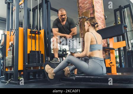 Test di fitness con personal trainer. Elegante ragazza caucasica skinny nella sua 20s e il suo maschio muscolare istruttore in palestra. Attrezzature professionali per l'esercizio. Foto di alta qualità Foto Stock