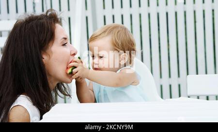 estate, in giardino, una divertente ragazza bionda di un anno tratta sua madre con anguria, la nutre dalle mani, la ragazza mangia anche anguria. Foto di alta qualità Foto Stock