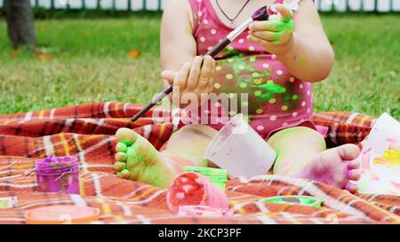 un bambino piccolo, una ragazza di un anno che gioca, dipinge con le pitture delle dita, si adorna, in giardino, seduta su una coperta, copriletto, su erba, prato, in estate. Foto di alta qualità Foto Stock