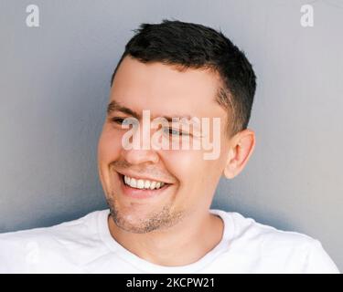 Ritratto di un uomo divertente con un sorriso stoppia su uno sfondo chiaro. Un ragazzo emozionale in una T-shirt bianca ride guardando via. Buon umore, cordiale Foto Stock