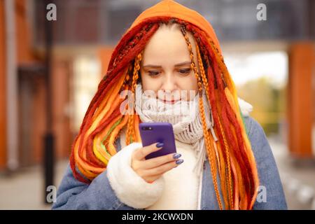 Giovane donna con capelli luminosi in abiti caldi che naviga sul telefono cellulare, in piedi sulla strada cittadina Foto Stock