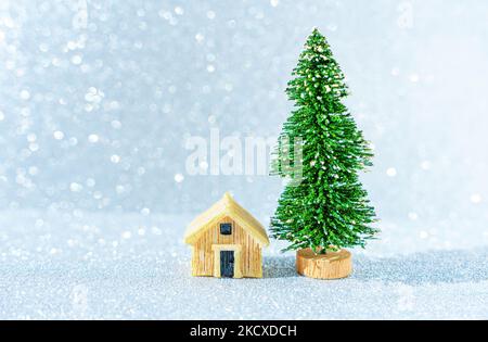 Modello di casa in miniatura posto da un albero di Natale giocattolo su uno sfondo scintillante. Foto Stock
