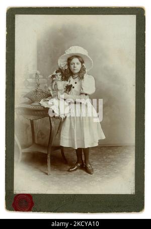 Edwardian era studio ritratto armadio scheda di bella ragazza guardando da una famiglia privilegiata, classe superiore / media indossando un grande cappello bianco, bel vestito bianco (partito ?) con maniche gonfie, guanti e collant bianchi e tenendo la bambola in cina. Forse una fotografia di compleanno. Sul retro è scritto Clara R. J. Whant, di 11 1909 anni. Dallo studio di Edward Sharp Islington e Westminster Bridge London, Regno Unito, datato 1909. Foto Stock