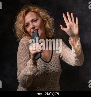 Una donna bionda simula il canto di un concerto utilizzando un telecomando come microfono Foto Stock