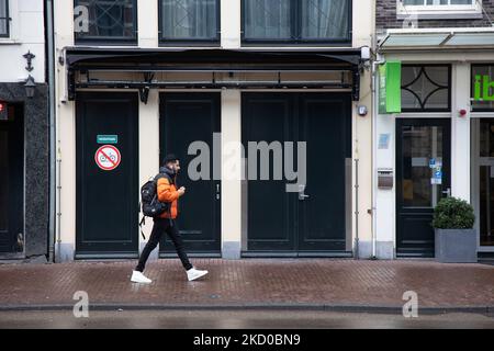 Vita quotidiana ad Amsterdam con persone che passano di fronte a caffè, bar e ristoranti chiusi. Locali e alcuni turisti nelle tranquille strade di Amsterdam durante il blocco nella capitale olandese con negozi e negozi che appaiono con chiuso con la tapparella metallica giù, caffè, bar e ristoranti anche chiusi con tavoli e sedie delle terrazze chiuse. I Paesi Bassi sono stati la prima nazione europea a dichiarare il blocco completo per combattere la nuova variante di Omicron che si surge. Dopo un improvviso ordine governativo prima di Natale, il paese ha chiuso tutti i negozi non essenziali, caffè, ristoranti, ba Foto Stock